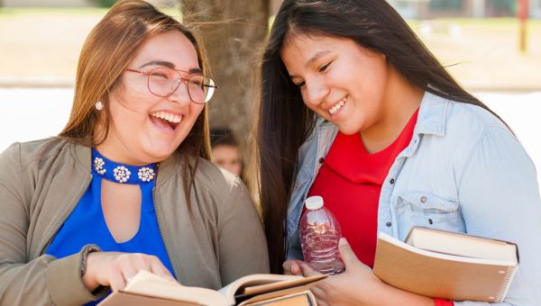 Two young people standing under a tree looking at the same page from an open textbook