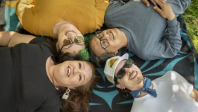 Four Indigenous people of varying ages and genders lie in a circle outdoors on a blanket with their heads together. 