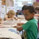 A classroom scene with young students sitting at a table, working on arts and crafts projects. A boy wearing glasses and a green shirt is in the foreground, focused on his work.