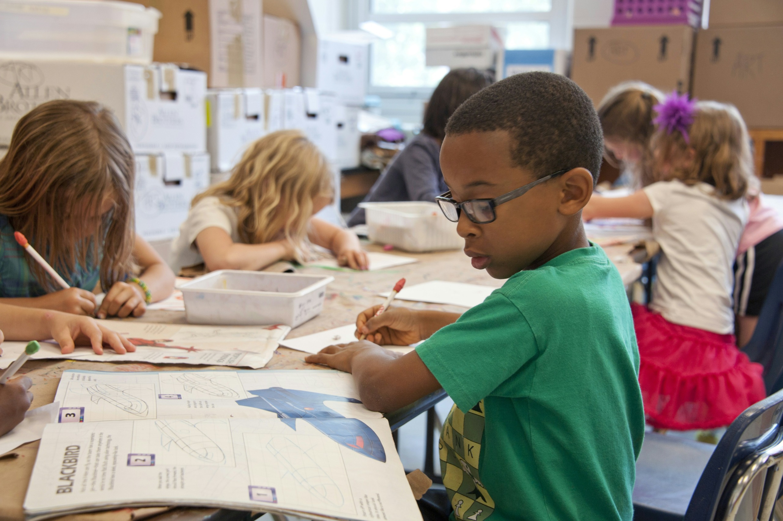 A classroom scene with young students sitting at a table, working on arts and crafts projects. A boy wearing glasses and a green shirt is in the foreground, focused on his work.