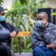 Two individuals sitting on a bench outdoors, both wearing face masks. The person on the left wears a dark coat, a knitted hat with a pom-pom, and sunglasses, while the person on the right wears a patterned sweater and rests their hand on their cheek. The background features lush greenery and a blurred figure in the distance. Photo by Centre for Ageing Better