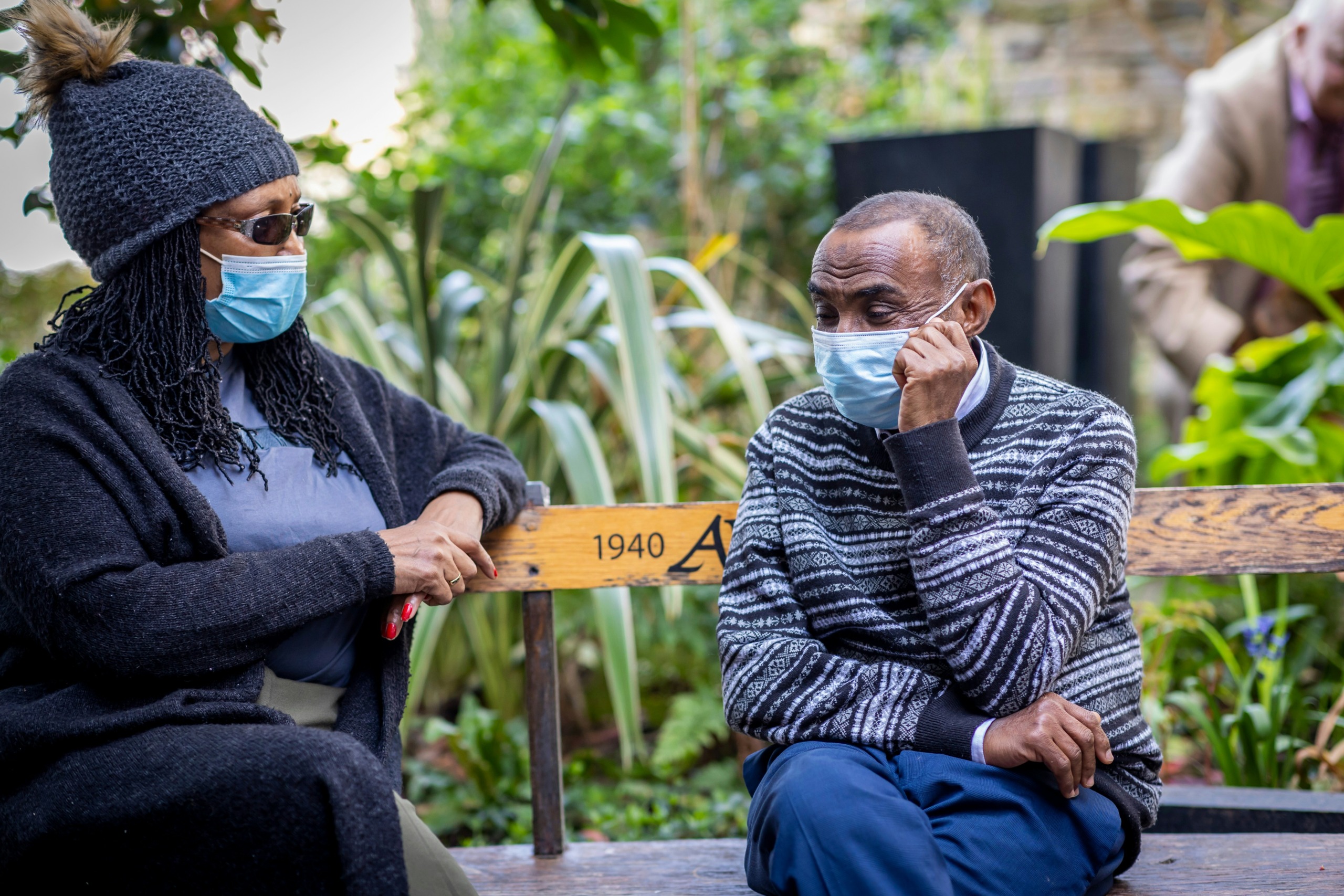 Two individuals sitting on a bench outdoors, both wearing face masks. The person on the left wears a dark coat, a knitted hat with a pom-pom, and sunglasses, while the person on the right wears a patterned sweater and rests their hand on their cheek. The background features lush greenery and a blurred figure in the distance. Photo by Centre for Ageing Better