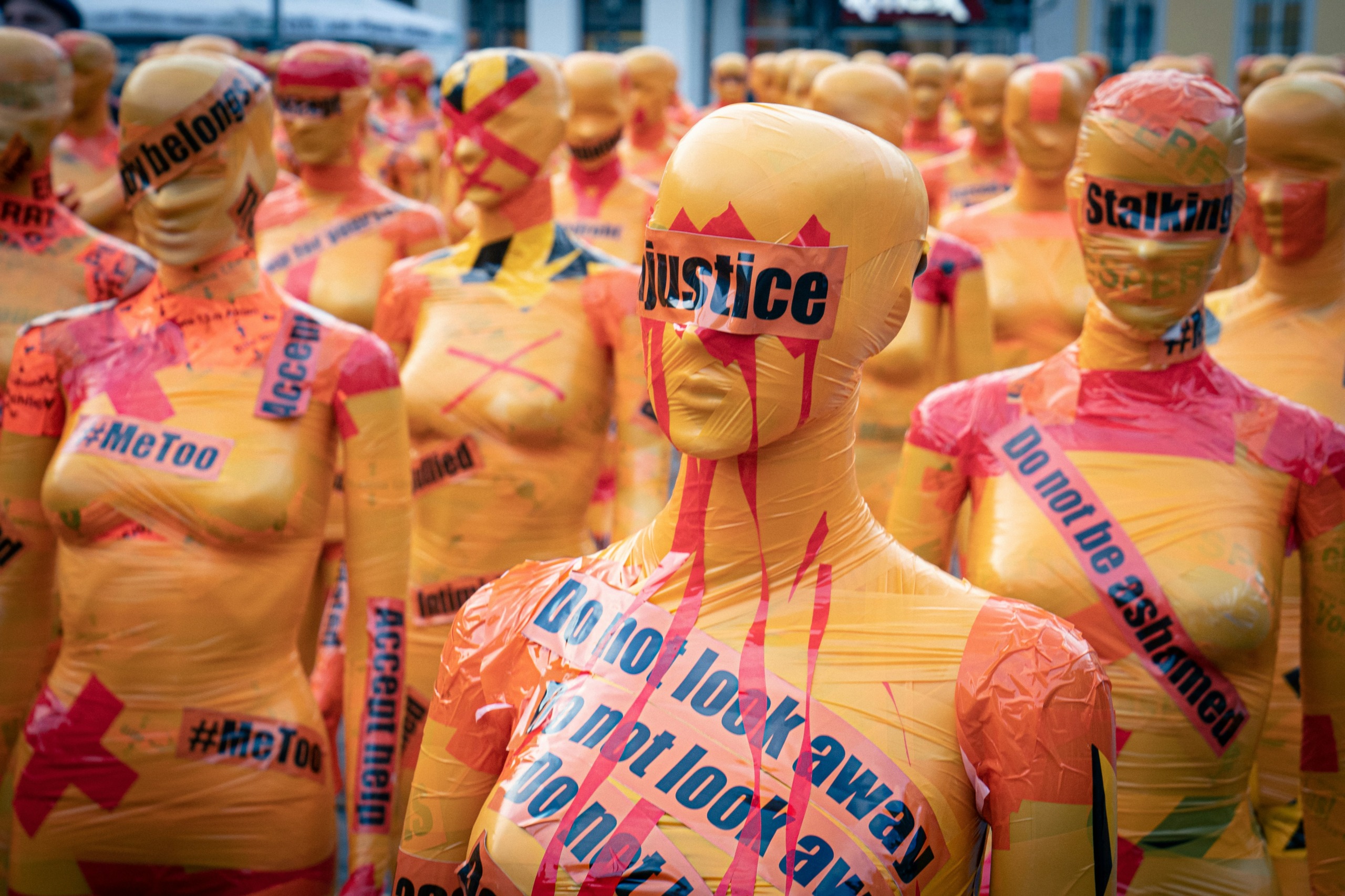 Dozens of mannequins wrapped in orange tape with protest messages such as "Justice," "#MeToo," and "Do not look away" displayed prominently. The installation highlights issues such as violence, stalking, and harassment, with bold text and symbols creating a powerful visual statement. Photo by Mika Baumeister