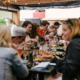 A group of people gathered around a long table in an outdoor setting, enjoying food and drinks. The table is filled with plates of food, glasses of beverages, and various items, creating a lively and social atmosphere. String lights hang above, adding a warm and inviting ambiance to the scene. Photo by Priscilla Du Preez