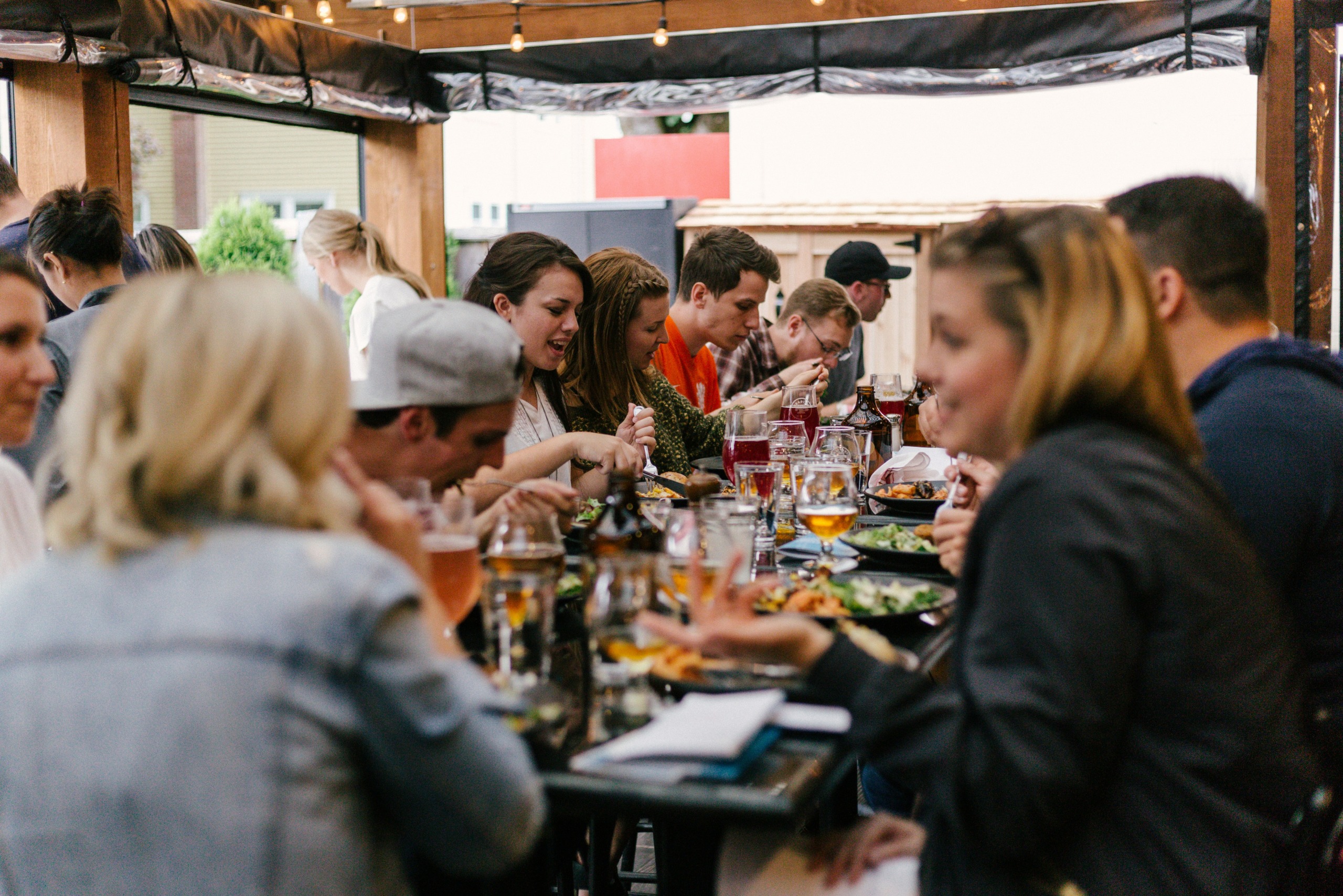 A group of people gathered around a long table in an outdoor setting, enjoying food and drinks. The table is filled with plates of food, glasses of beverages, and various items, creating a lively and social atmosphere. String lights hang above, adding a warm and inviting ambiance to the scene. Photo by Priscilla Du Preez