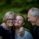 A happy family portrait featuring a young man with Down syndrome standing between an older man and woman. The woman, wearing glasses and a navy top, hugs the young man with a big smile. The young man is smiling warmly, and the older man looks at them affectionately, all against a soft green, natural background. photo by Nathan Anderson