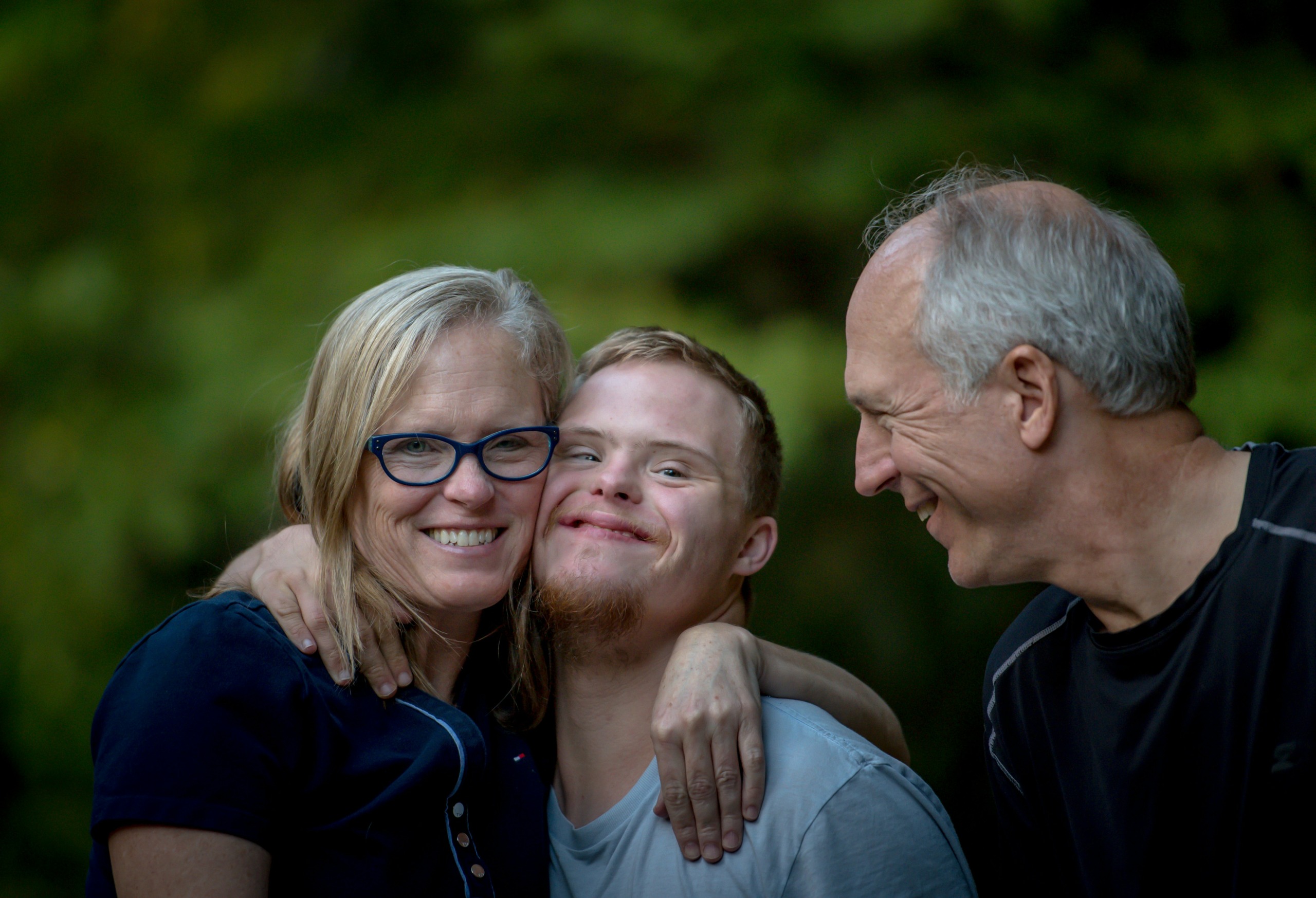 A happy family portrait featuring a young man with Down syndrome standing between an older man and woman. The woman, wearing glasses and a navy top, hugs the young man with a big smile. The young man is smiling warmly, and the older man looks at them affectionately, all against a soft green, natural background. photo by Nathan Anderson